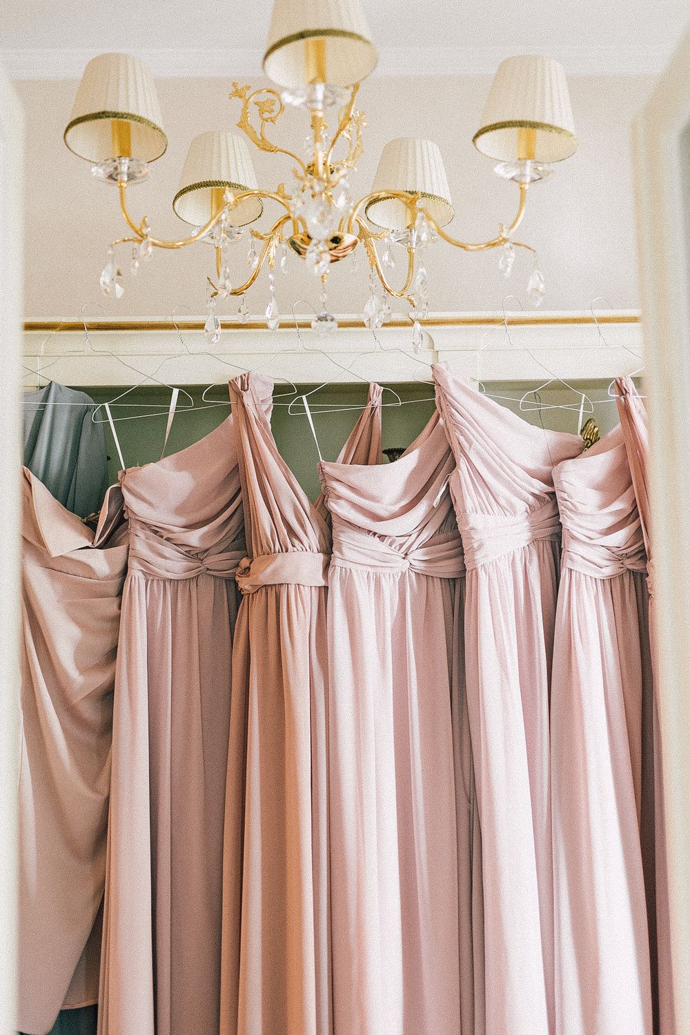 Blush pink bridesmaid dresses hanging on hangers beneath an elegant gold chandelier in a bridal suite.