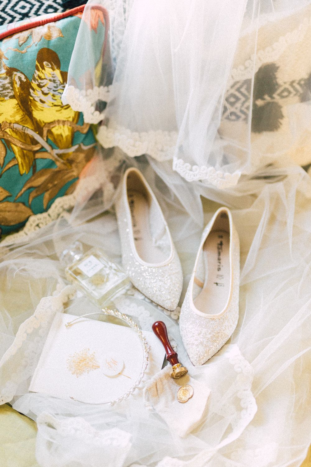 White lace bridal flats displayed on a veil with a wax seal, perfume bottle, and floral bag for a wedding flat lay.