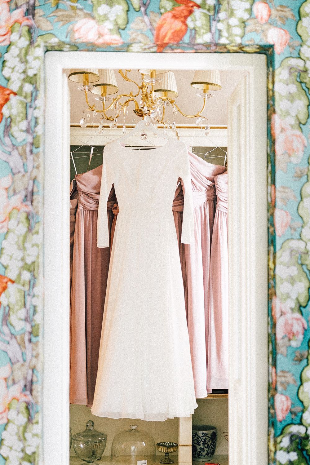 White wedding dress hanging in doorway with pink bridesmaid dresses and gold chandelier, framed by floral wallpaper.