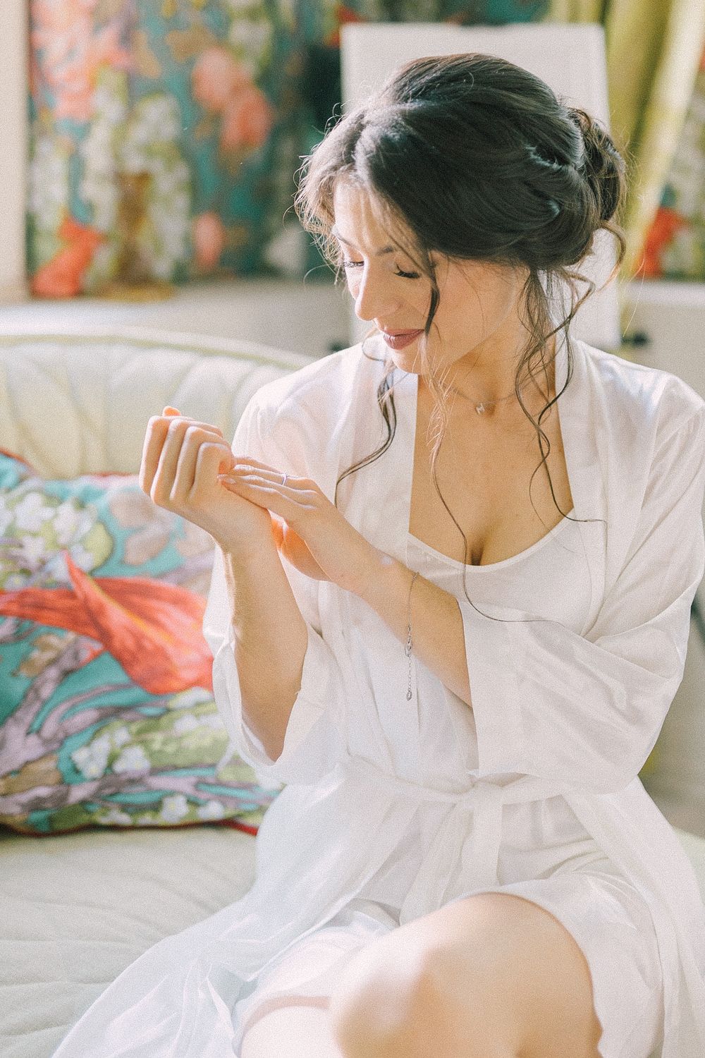 Dark-haired woman in white robe applying lotion to her hands while sitting on a cream sofa.
