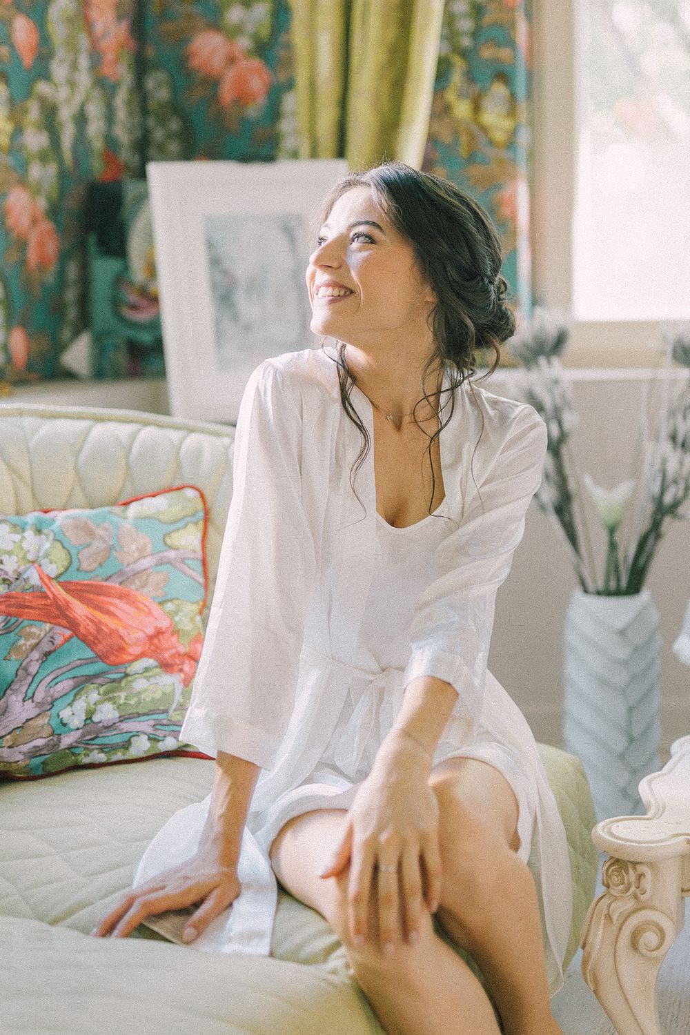 Smiling brunette woman in white robe sitting on cream sofa in bright floral decorated room.