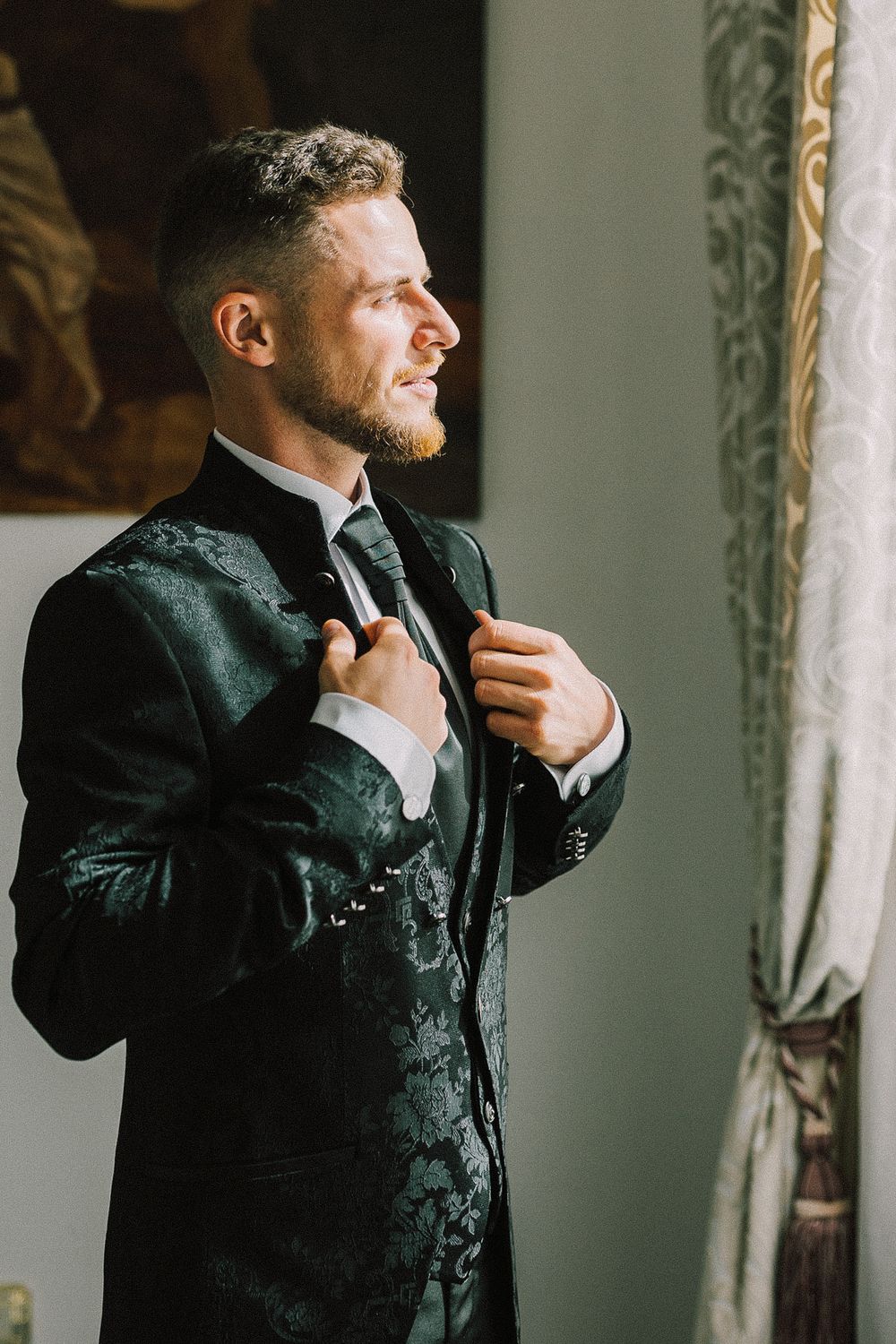A well-dressed groom in a dark floral suit adjusts his tie near a window, smiling confidently before his wedding.