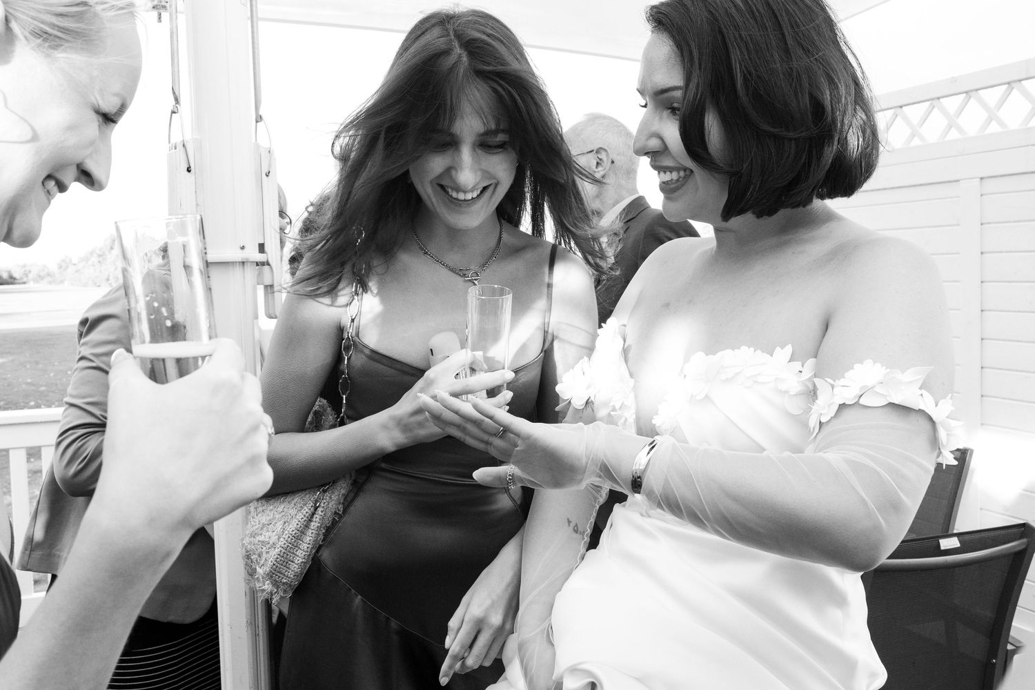 Black and white photo of a bride in white dress showing her engagement ring to two smiling women at an outdoor wedding celebration.