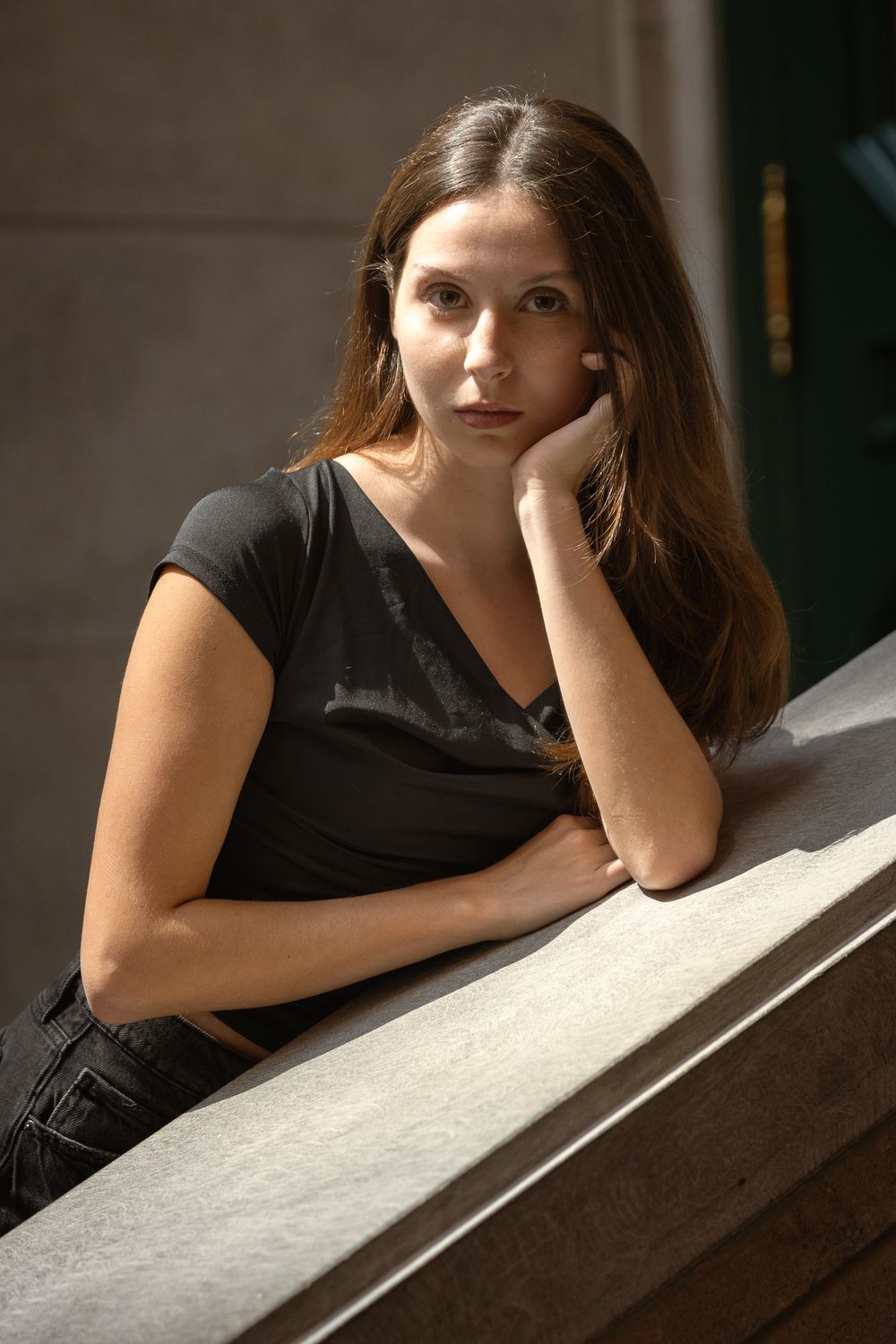 Jeune femme aux longs cheveux châtains accoudée à une rampe en pierre, la joue dans la main, en lumière naturelle