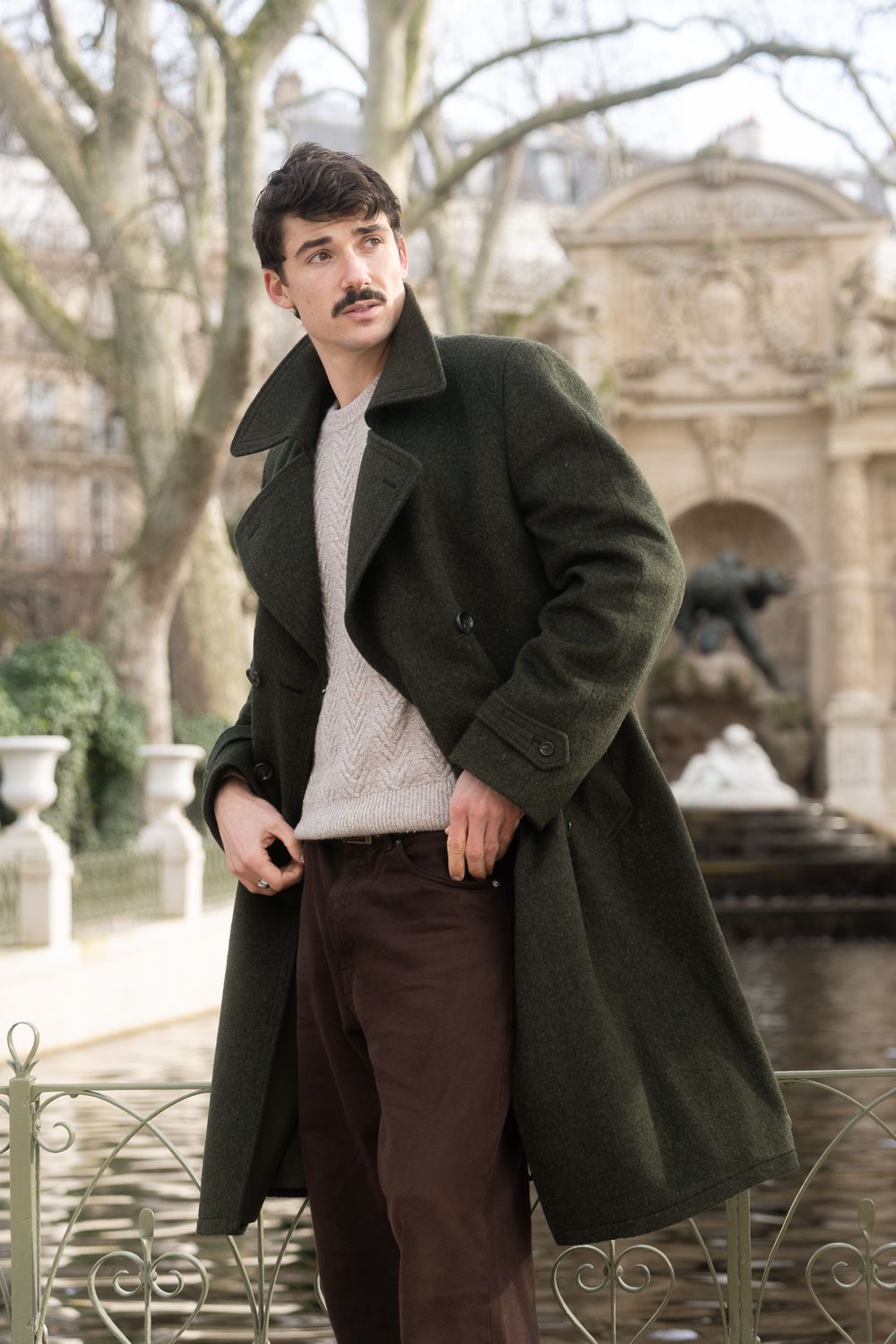 Homme à moustache en long manteau vert croisé et pull beige, la main dans la poche, devant un bassin et une fontaine