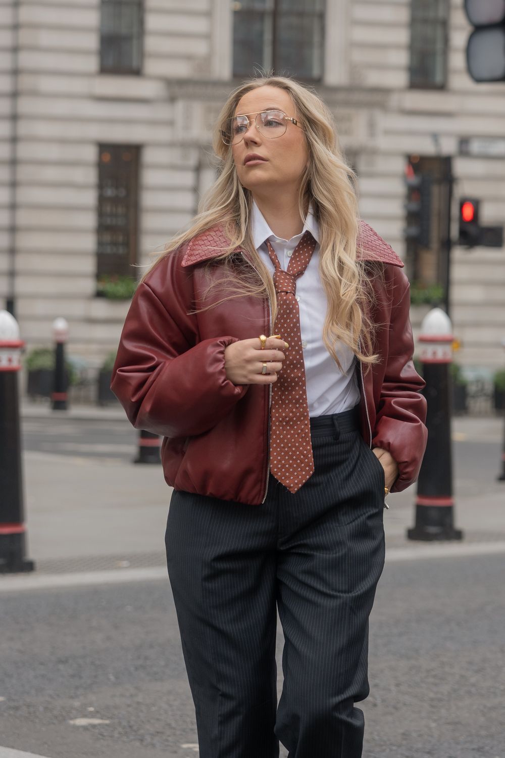 Femme blonde aux cheveux longs, lunettes aviateur, blouson en cuir bordeaux, cravate à pois et pantalon à rayures dans la rue