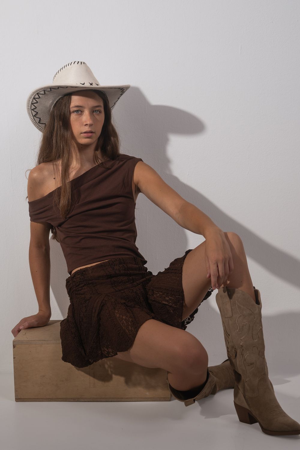 Jeune femme aux cheveux longs, chapeau de cowboy blanc et bottes beige, assise sur une caisse en bois en studio