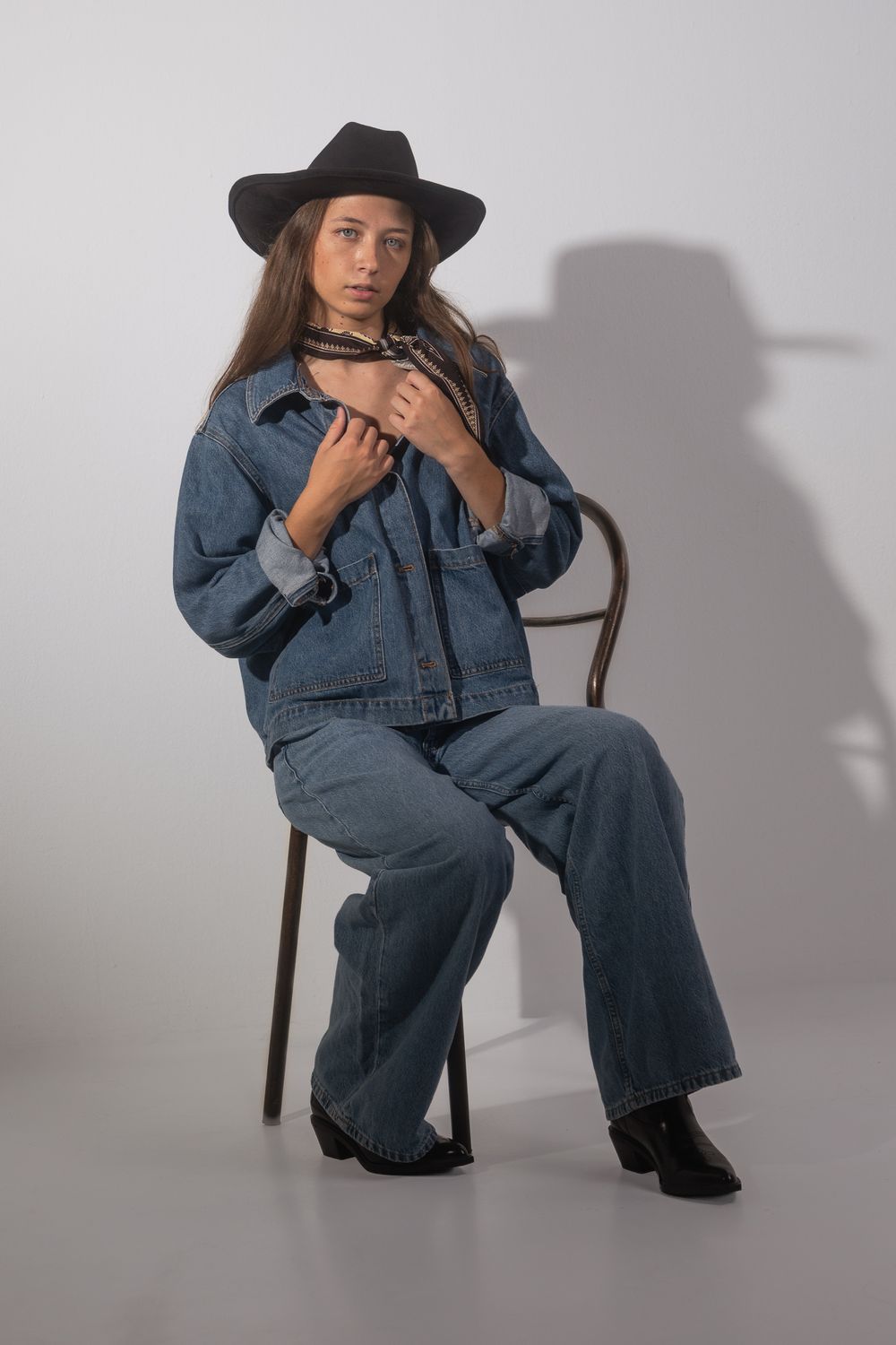 Jeune femme assise sur une chaise en bois courbé, chapeau noir, veste et jean en denim, foulard, fond studio gris