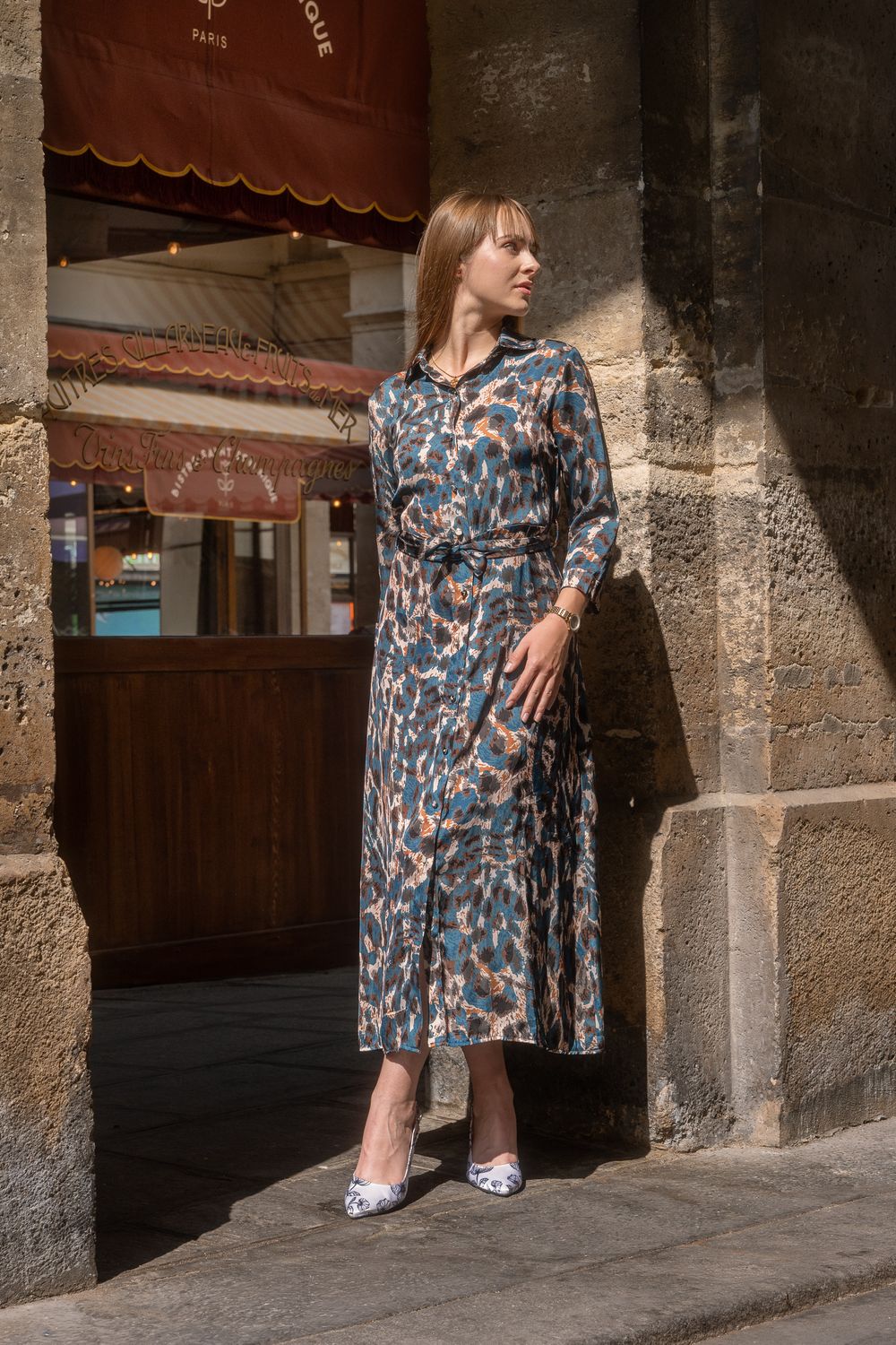 Femme en robe-chemise longue imprimé léopard bleu et marron, debout contre un mur de pierre, regardant au loin