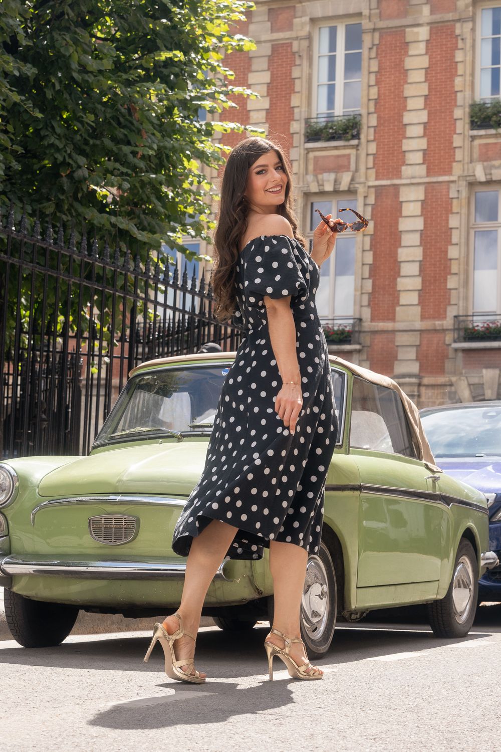 Jeune femme en robe noire à pois blancs, lunettes à la main, souriante devant une voiture ancienne verte