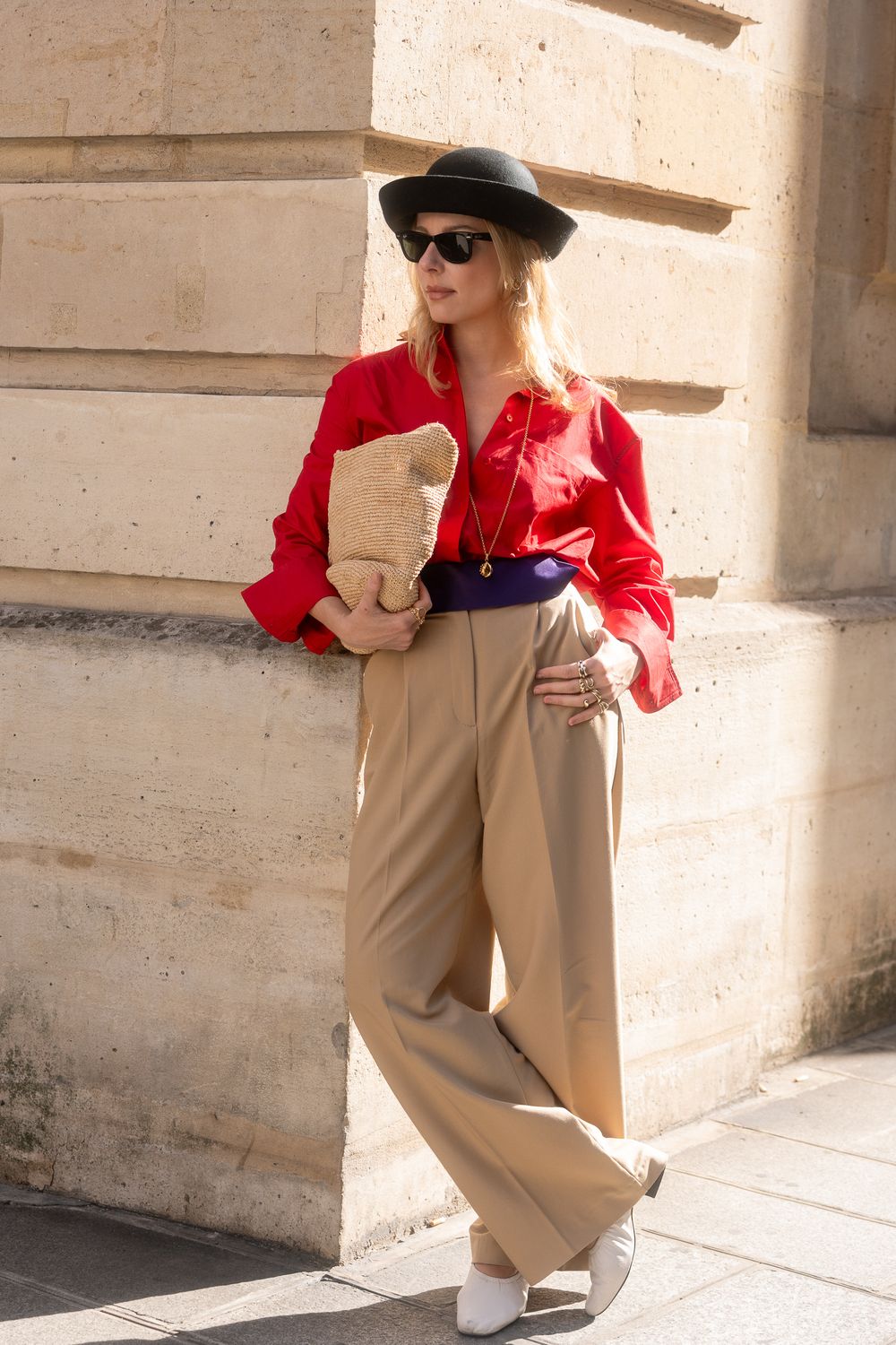 Femme blonde en chemise rouge, pantalon beige ample et chapeau noir, adossée à un mur en pierre claire