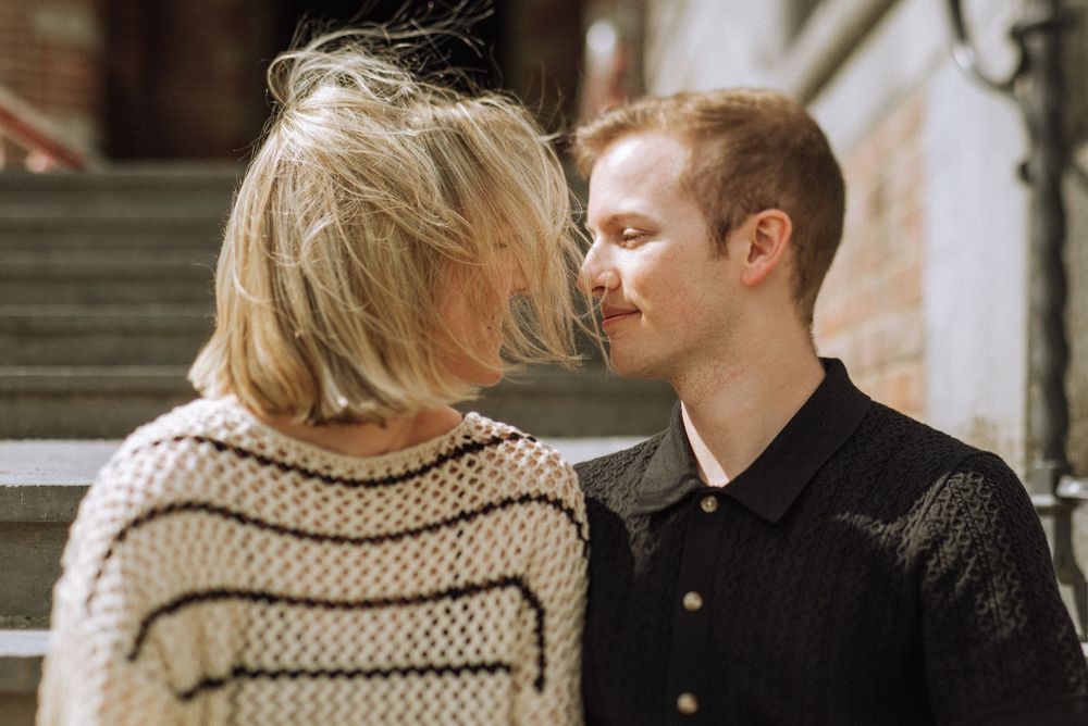 Couple sharing a romantic moment, nearly kissing outdoors on sunlit steps in casual stylish attire.