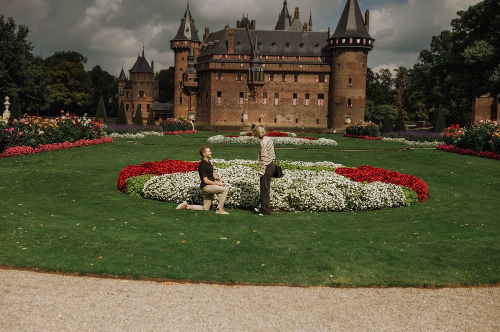Couple sitting in manicured gardens with red and white flowers before a grand medieval de Haar castle on a cloudy day.