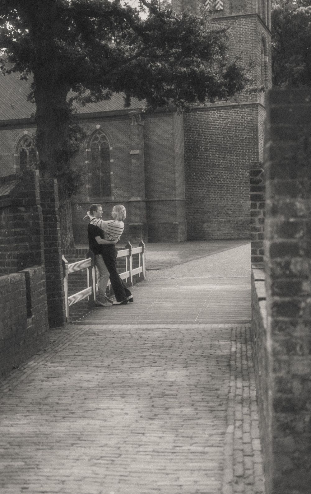 Black and white photo of a couple embracing on a cobblestone path beside a historic brick church. De Haar castle.
