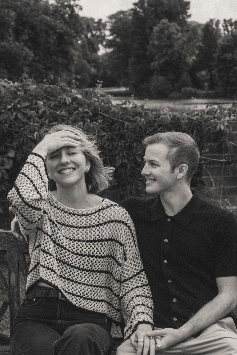 Black and white photo of a smiling couple sitting outdoors on a bench surrounded by lush green trees.