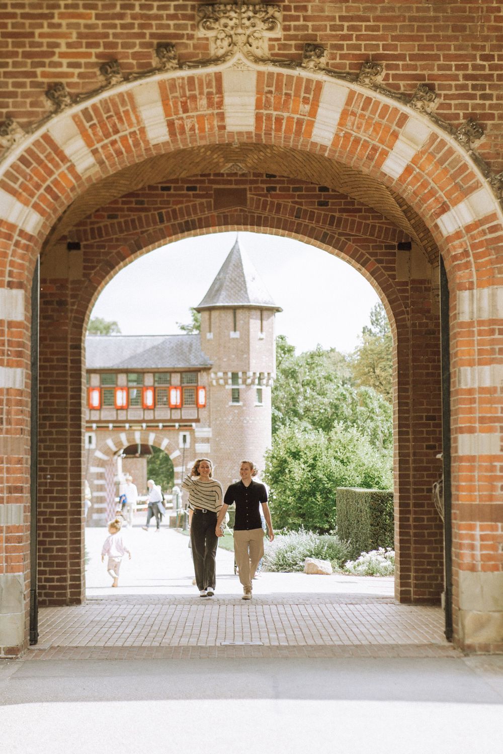 Two people walking through a red brick archway toward a historic building with a tower on a sunny day. De Haar castle