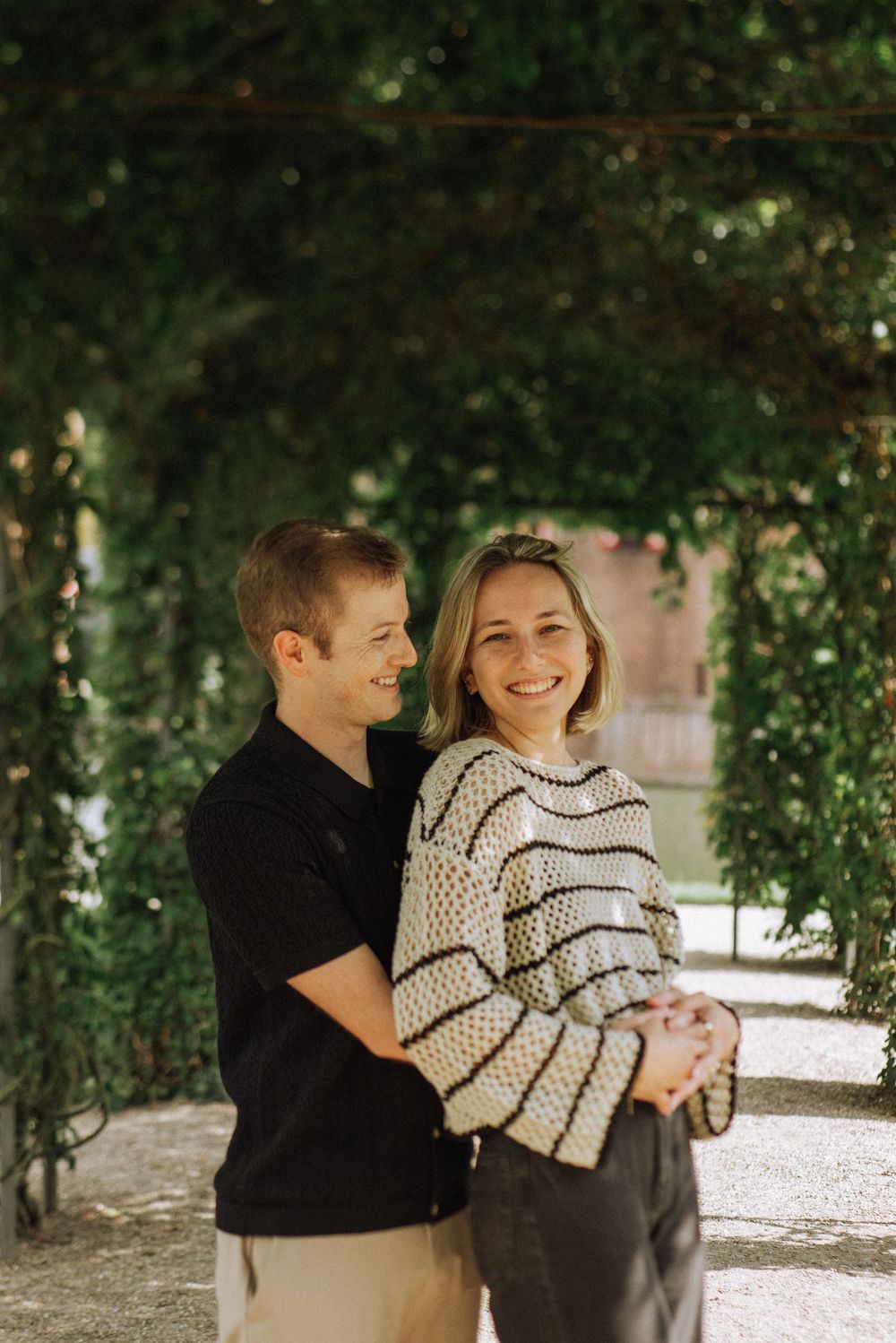 A smiling couple embraces under a lush green garden archway, the woman in a striped sweater looking at the camera.