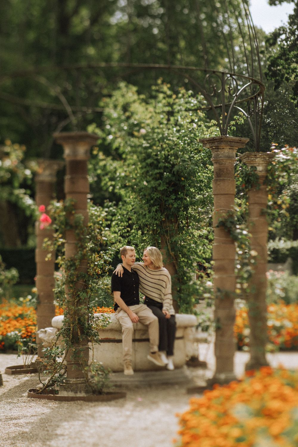 Couple sitting together under a stone pergola covered in vines in a beautiful garden with orange flowers.