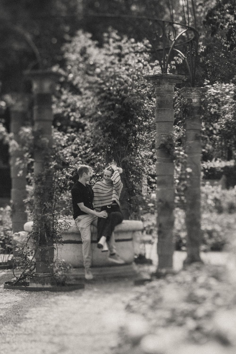 Black and white photo of a couple sitting together on a stone wall in a lush garden with ornate pillars.