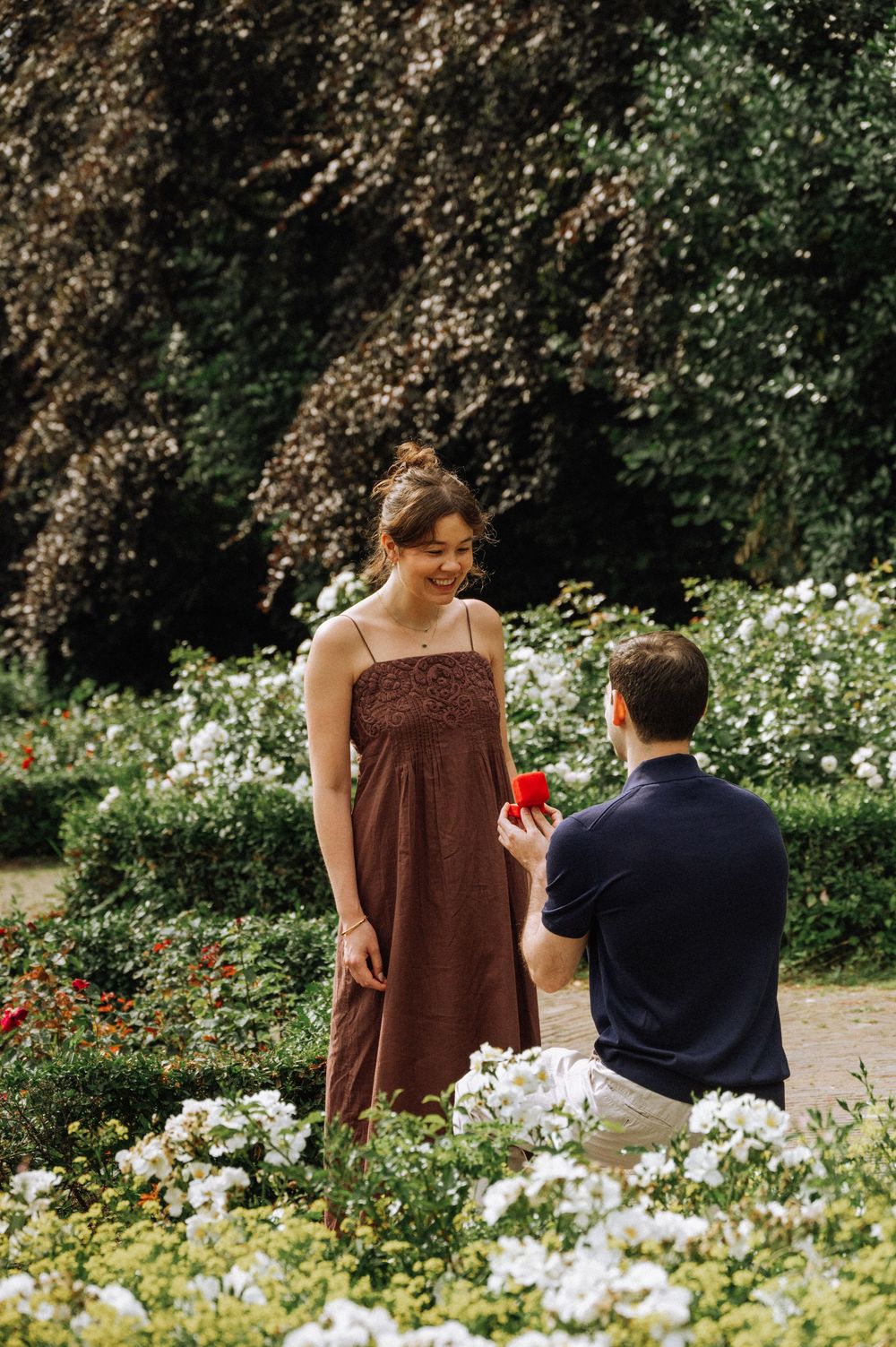 Man kneeling in a garden proposing to a smiling woman in a brown dress, holding a red ring box.
