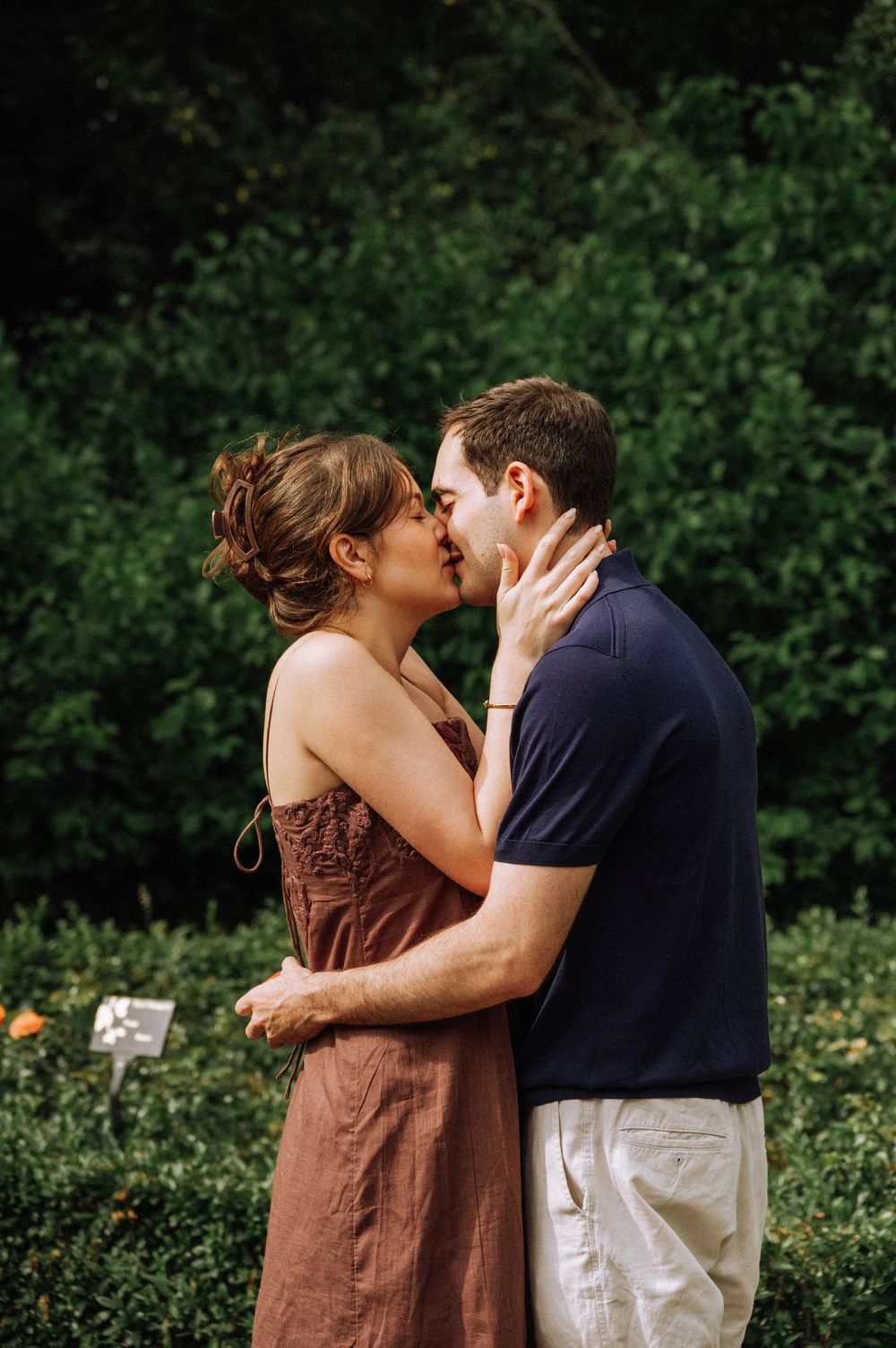 A couple shares a romantic kiss outdoors, surrounded by lush green hedges, the woman in a brown dress holding the mans face.