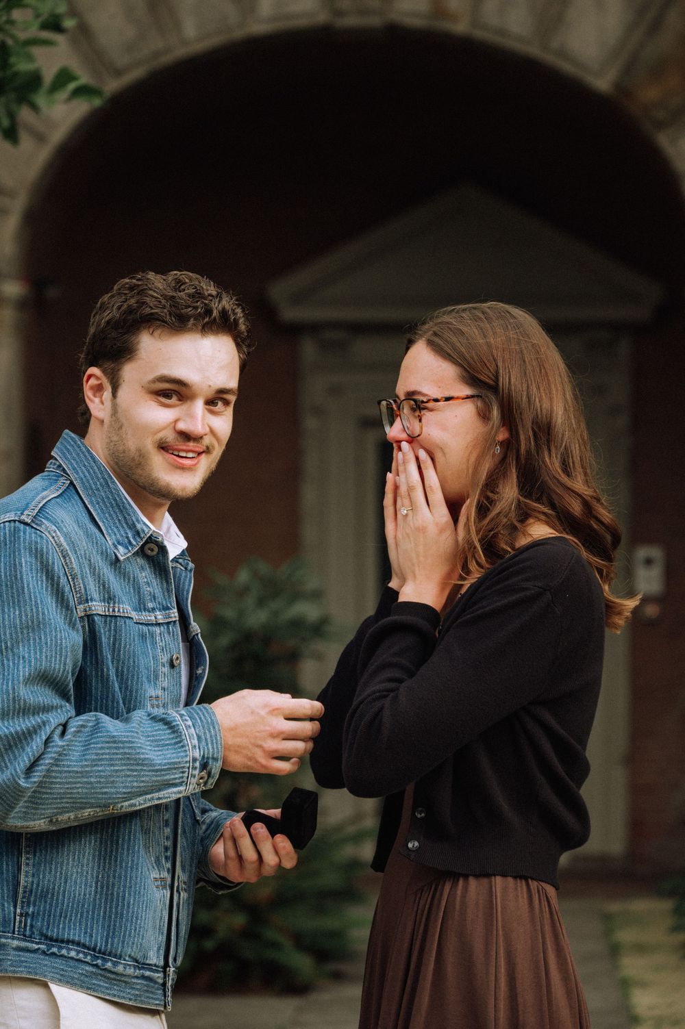Man in denim jacket holds ring box while woman covers her mouth in surprise during outdoor marriage proposal.