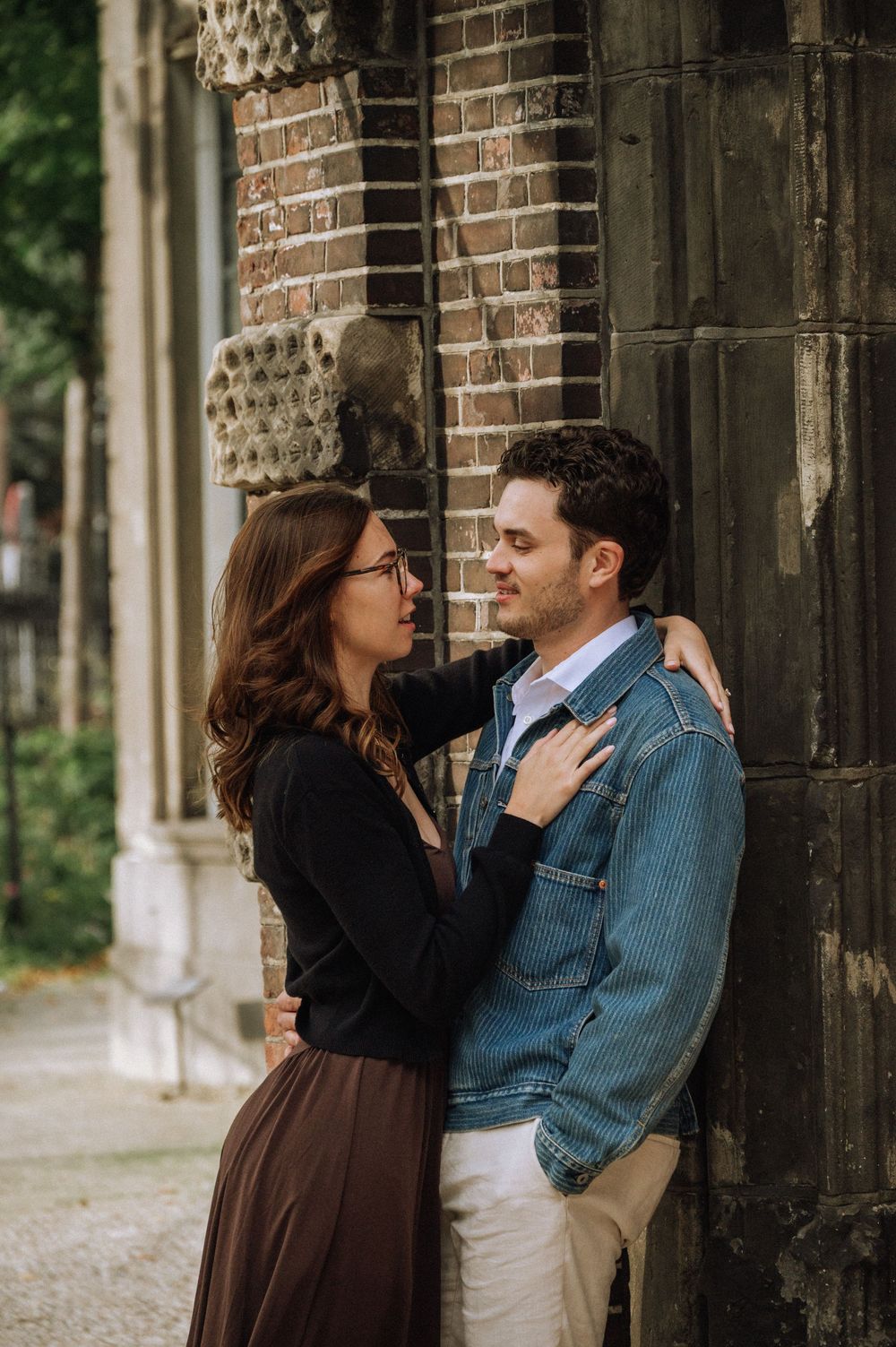 A couple shares a romantic moment leaning against a brick wall outdoors, the woman in glasses gazing at the man in a denim jacket.