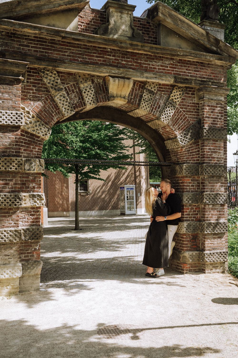 Couple embracing under a historic brick archway in a sunlit courtyard surrounded by lush green trees.