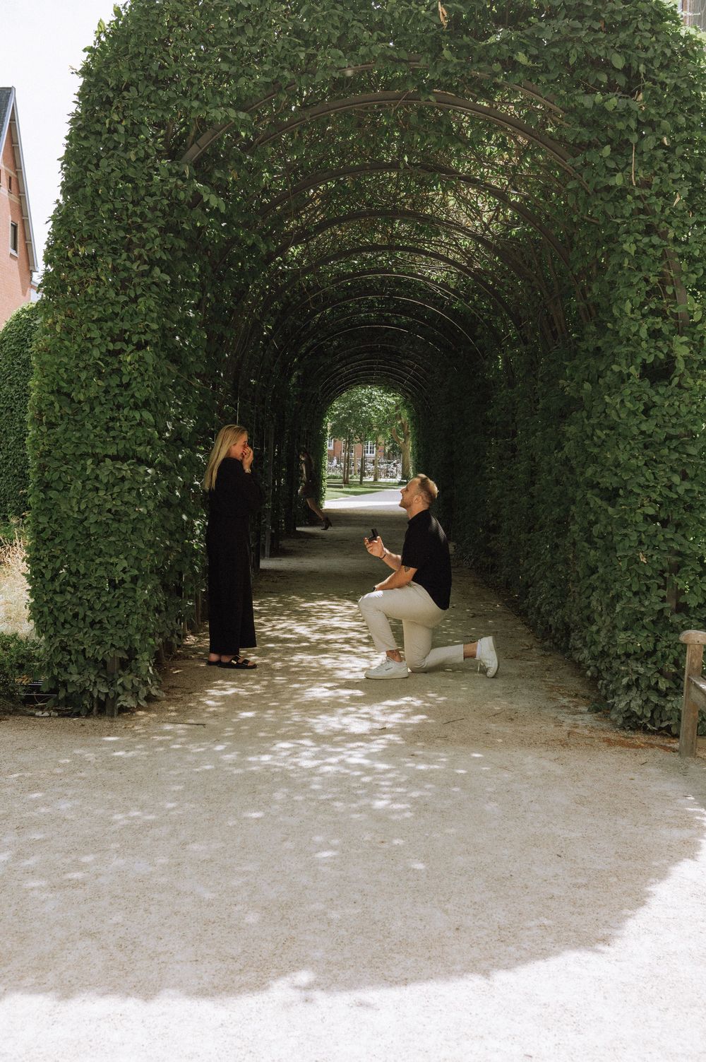 A person kneeling to propose in a lush green arched garden tunnel to their partner.