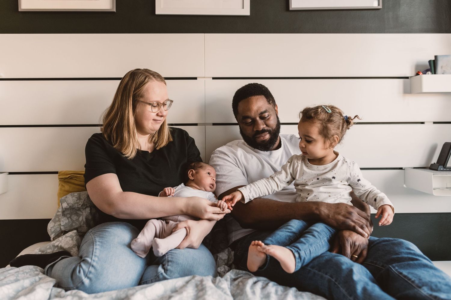Interracial family sitting on bed, parents holding newborn baby and toddler, smiling together in cozy bedroom.