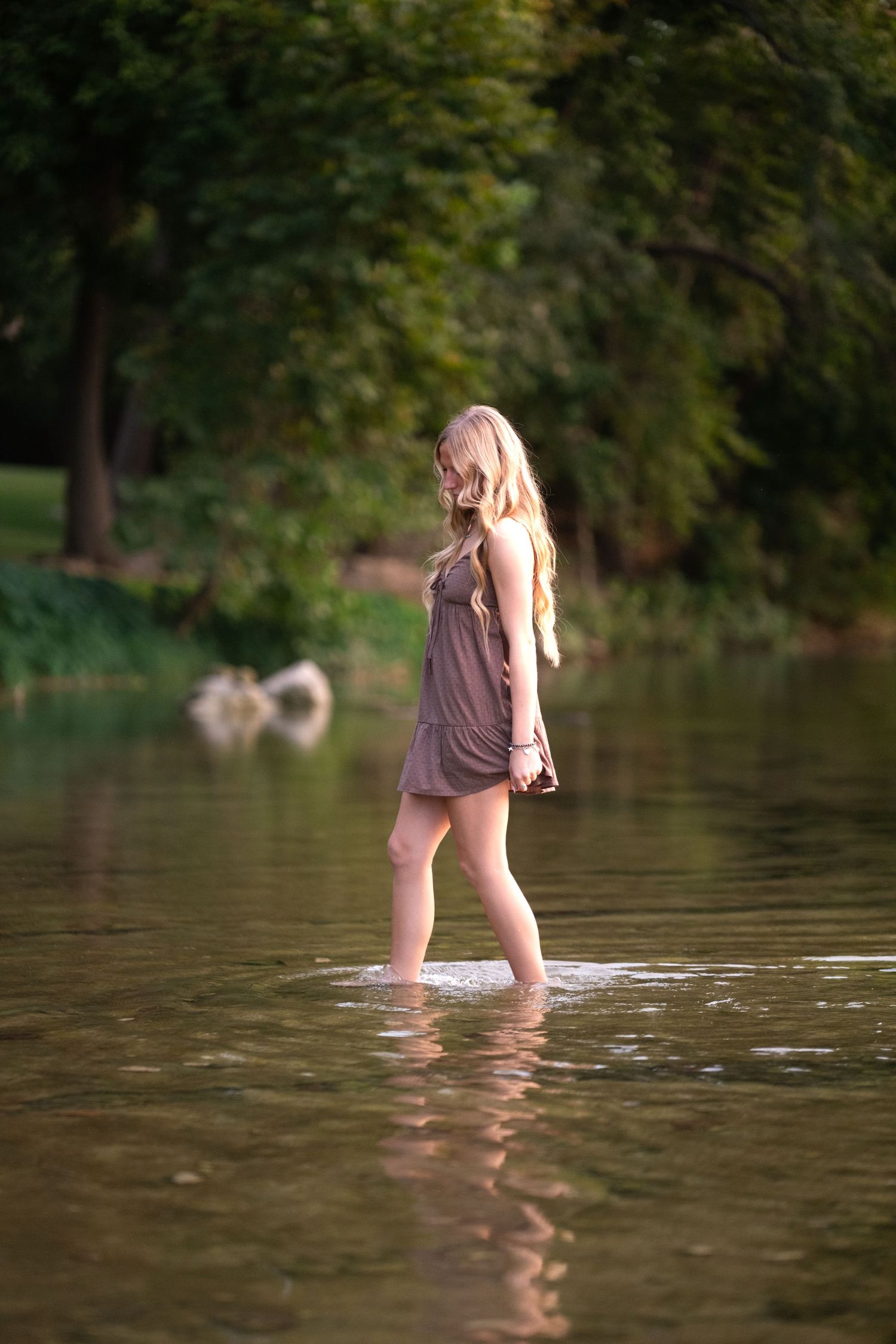 Young blonde woman in brown dress wading through calm river water surrounded by lush green trees.