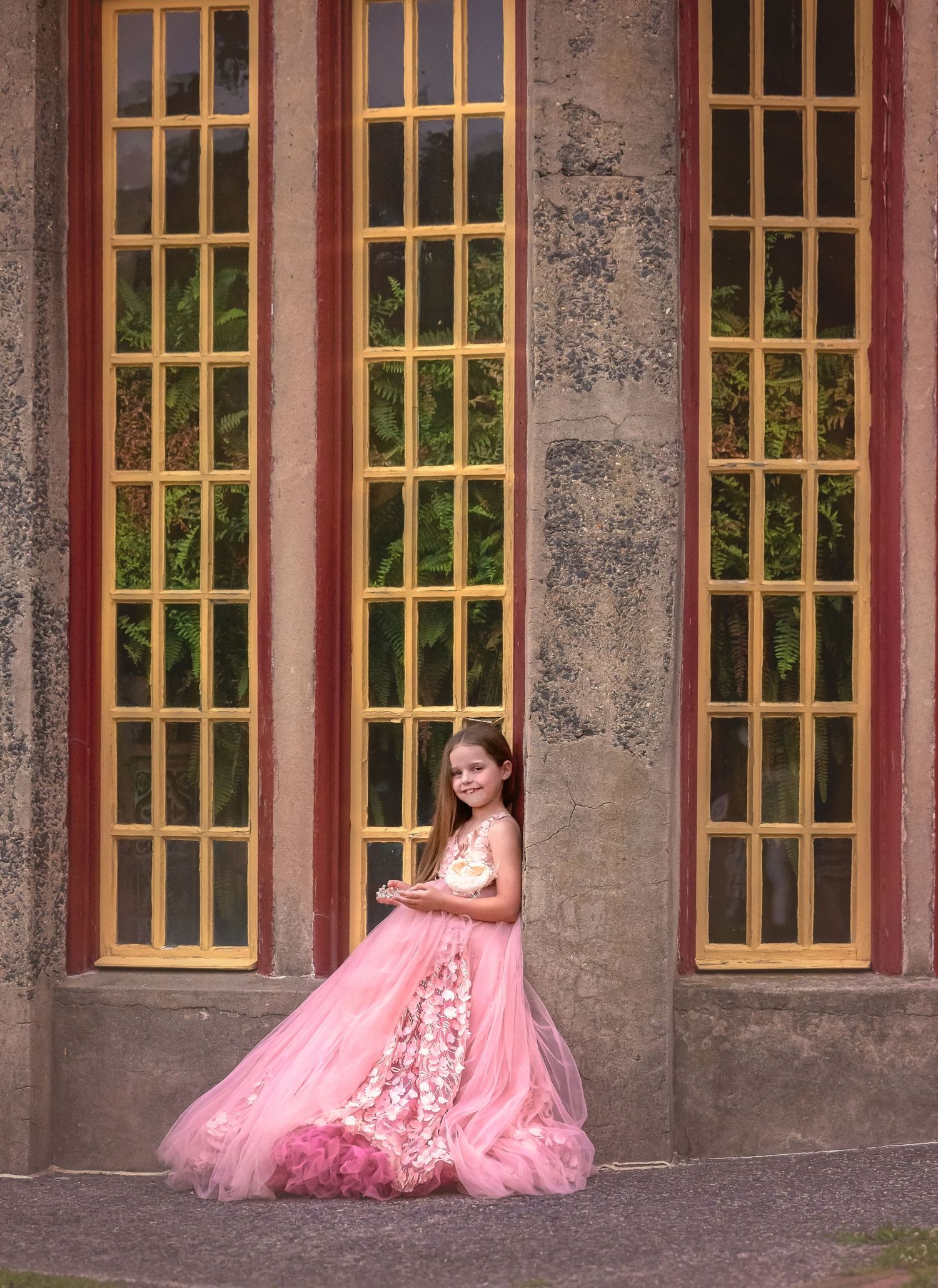 Young girl in pink princess gown poses gracefully against stone building with tall yellow-framed windows.