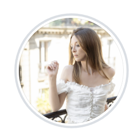 Woman with long brown hair in a white off-the-shoulder ruffled top stands on a wrought-iron balcony, looking to the side.