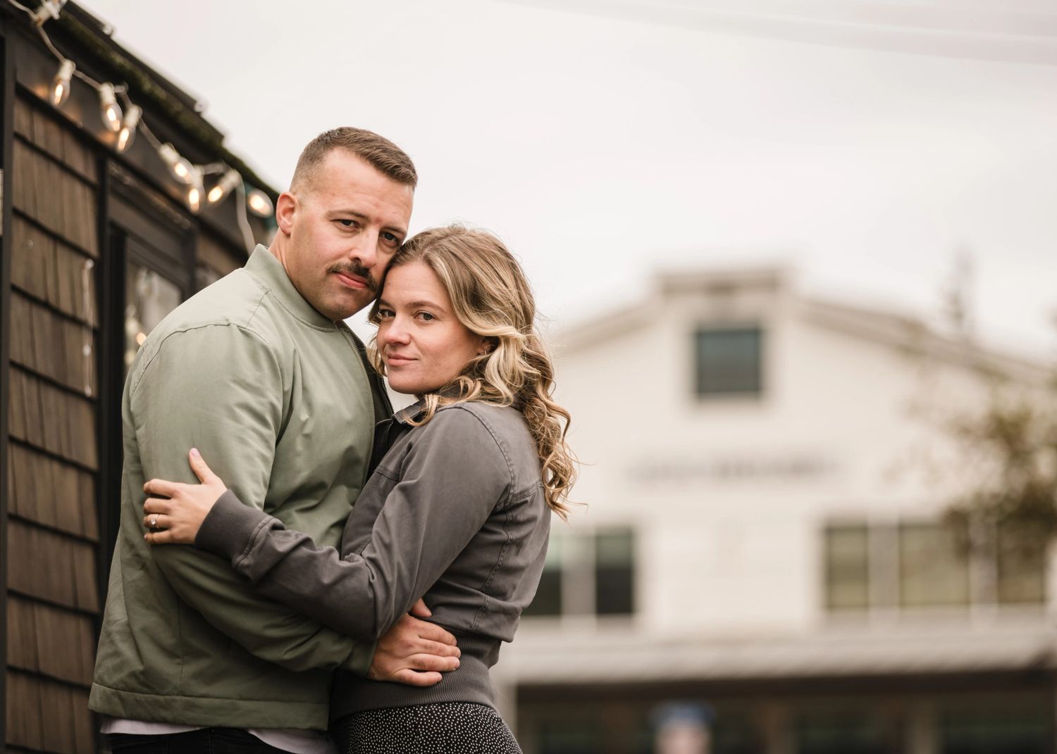 Couple embracing outdoors against a shingled wall, gazing toward camera with gentle affection. Engagement at Rockport Harbor, Rockport, MA