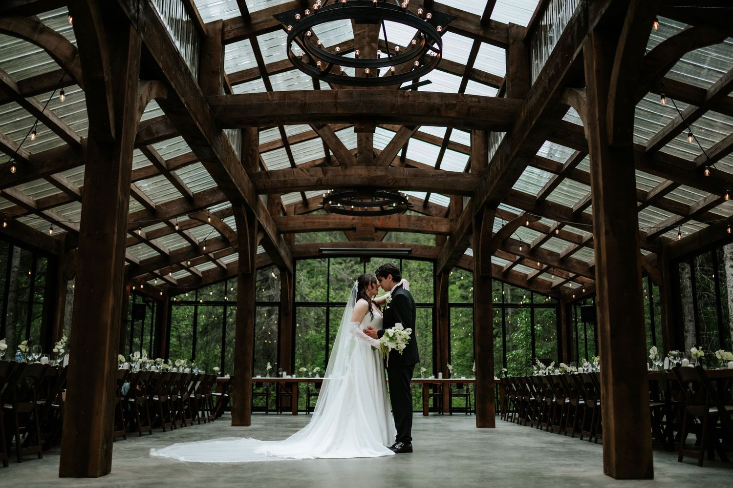 Bride and groom embrace in the center of an elegant timber-framed wedding venue with glass walls, wooden beams, and rows of chairs.