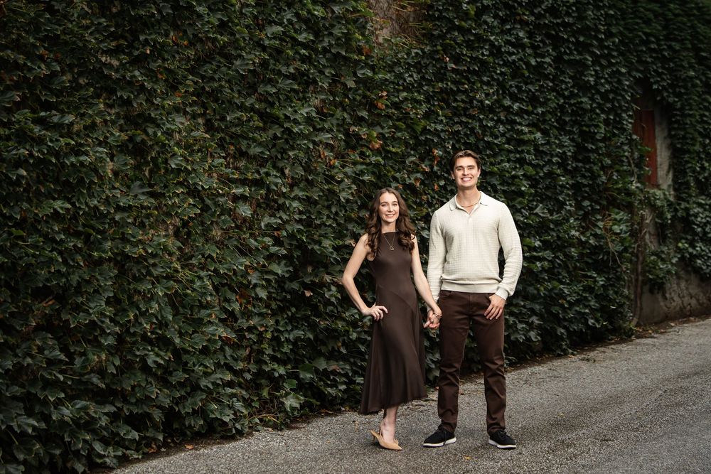 Smiling couple holding hands on a quiet alley path in front of a wall covered in lush green ivy during an engagement photo session.