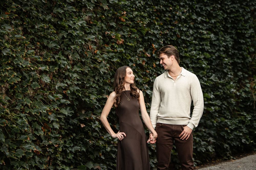 Smiling couple holding hands in front of a lush green ivy wall, the woman in a brown dress and the man in a cream sweater.
