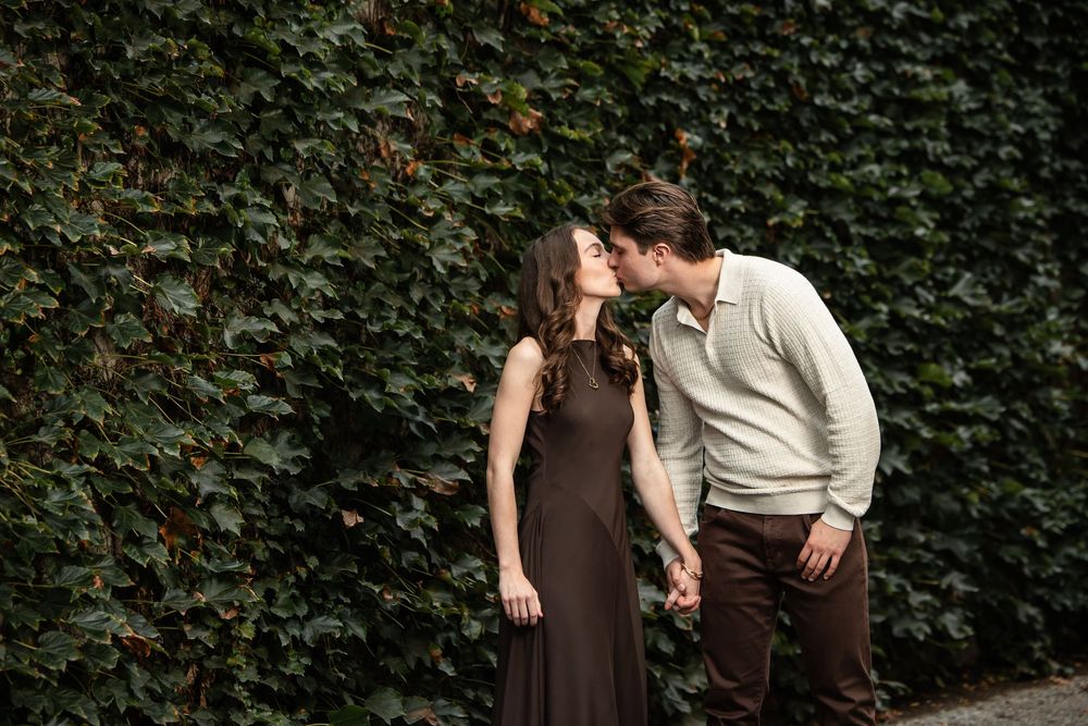 Couple in an olive dress and cream sweater share a kiss while holding hands in front of a lush green ivy wall.
