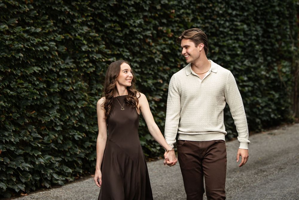 Smiling couple holding hands and walking along a path beside a lush green ivy wall, she in a brown dress and he in a cream sweater.