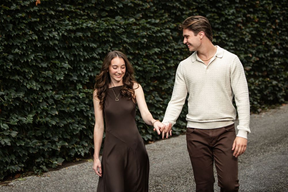 Smiling couple holding hands while walking along a paved path beside a dense green hedge, she in a brown dress and he in a cream sweater.