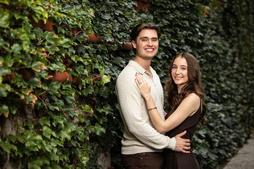 Smiling couple embracing in a romantic engagement-style portrait in front of a lush green ivy-covered wall.