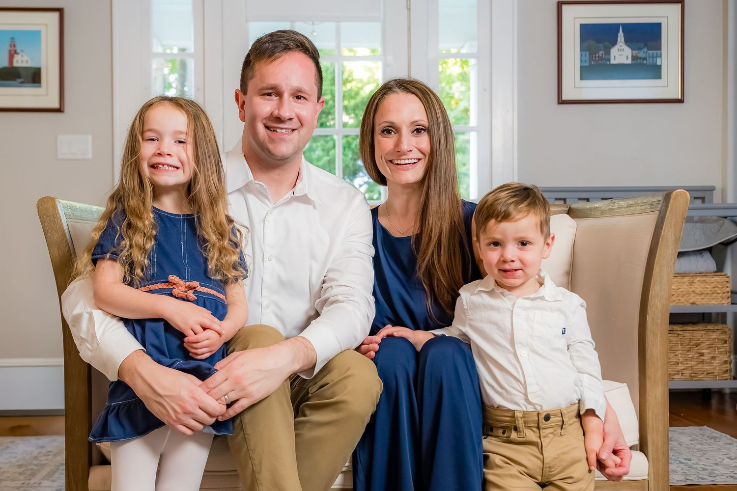 Smiling family of four posing on a cozy cushioned bench, with a young girl in a blue dress and a little boy in a white shirt and khakis.