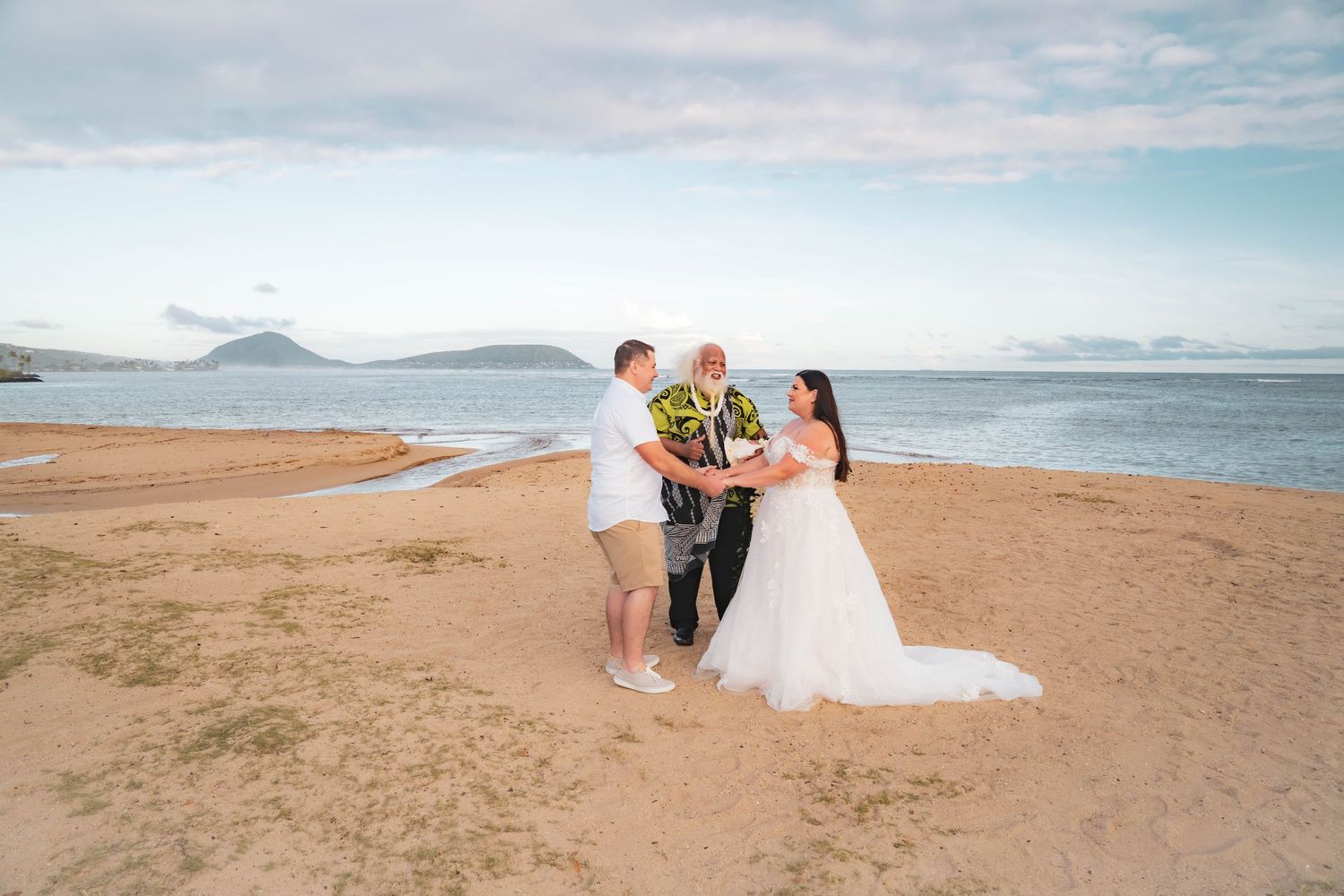 A bride in a white gown receives a flower lei from the groom during a beach wedding ceremony with an officiant by the ocean.