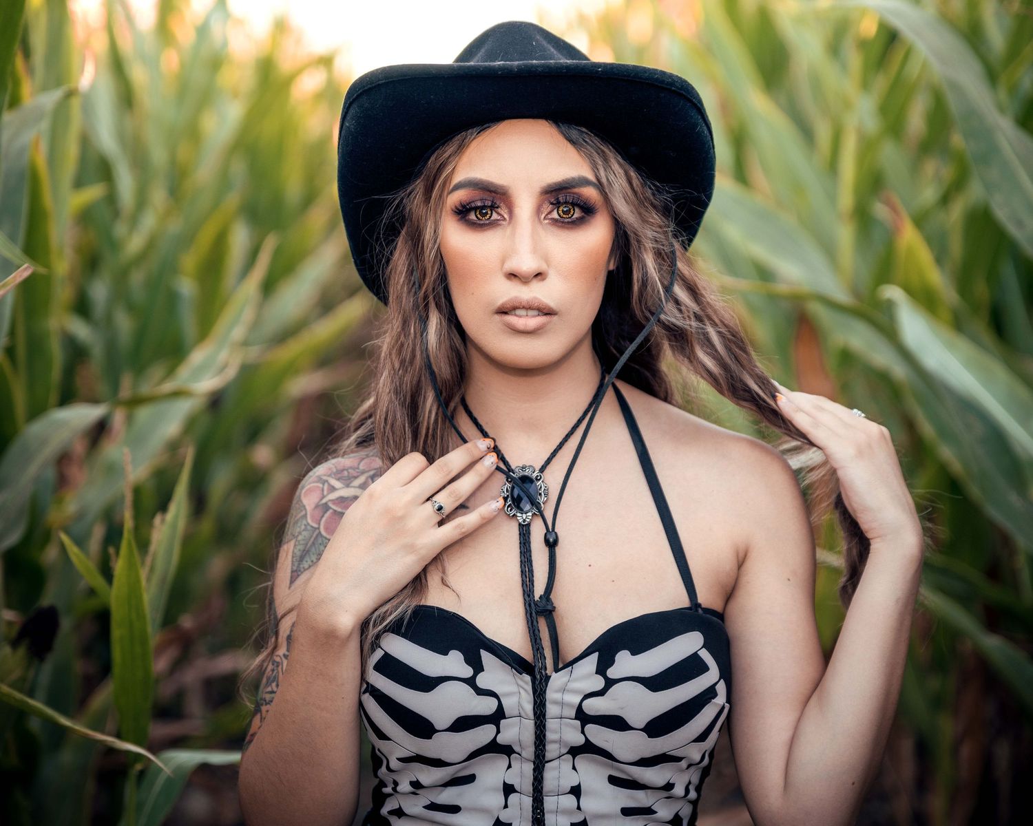 Female model in a cornfield at sunset wearing a cowboy hat and skeleton top, with tattoos and horror contacts, blending beauty and horror
