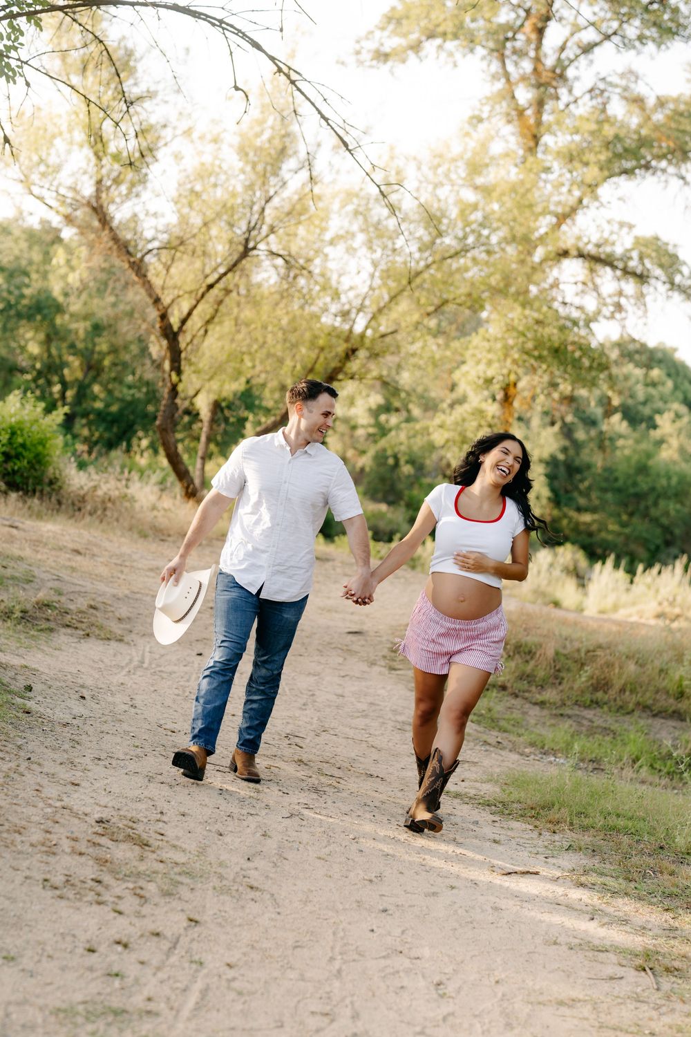 Happy pregnant couple holding hands and walking on a sunny dirt path surrounded by green trees.