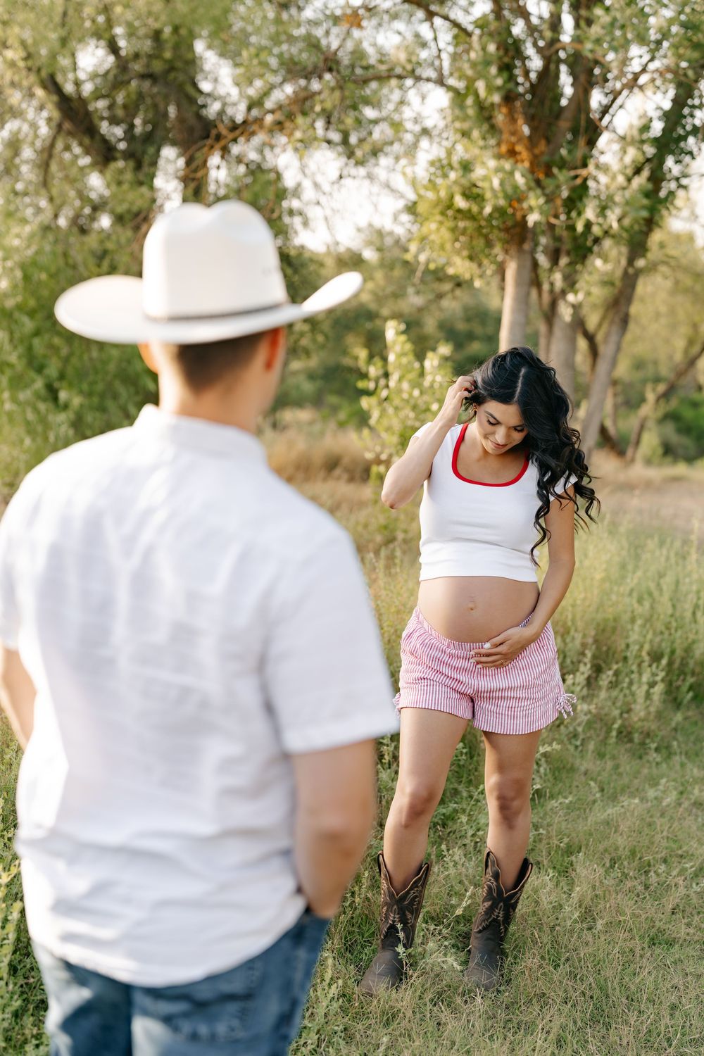 Pregnant woman in white crop top and pink shorts with cowboy boots stands in a field facing a man in a white hat.