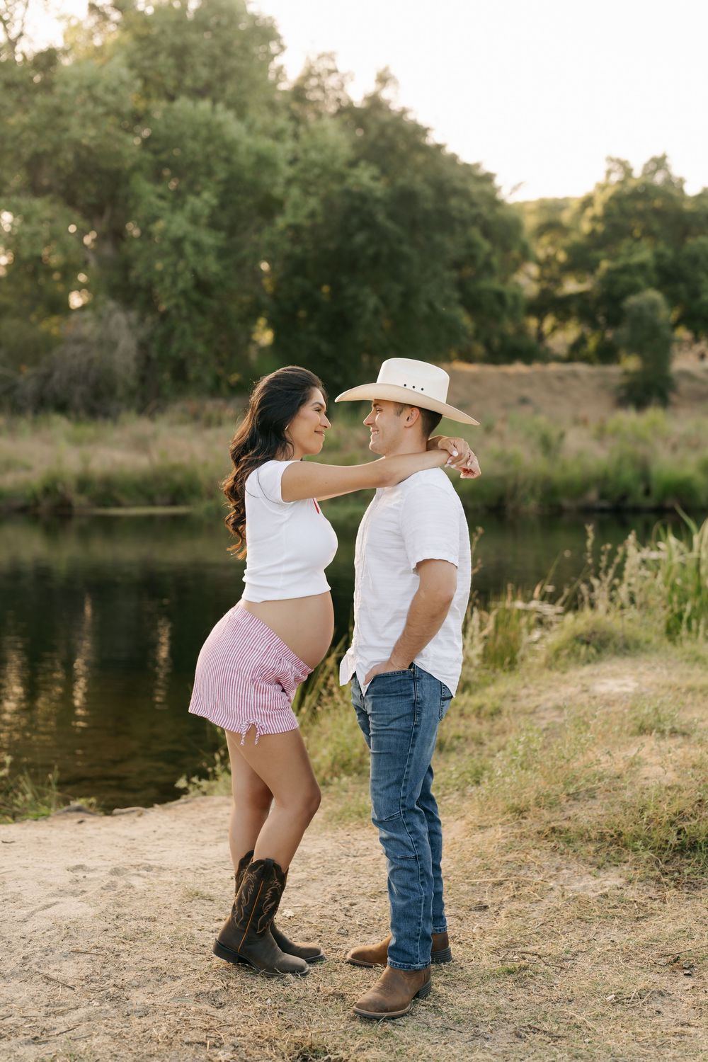 Pregnant couple in western wear posing by a pond, woman in white top and pink shorts, man in cowboy hat and jeans.