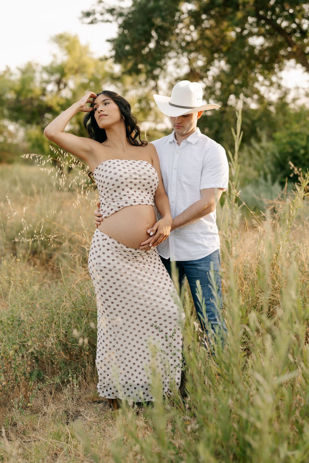 Pregnant couple in a field, woman in polka dot dress, man in white shirt and cowboy hat, embracing outdoors.
