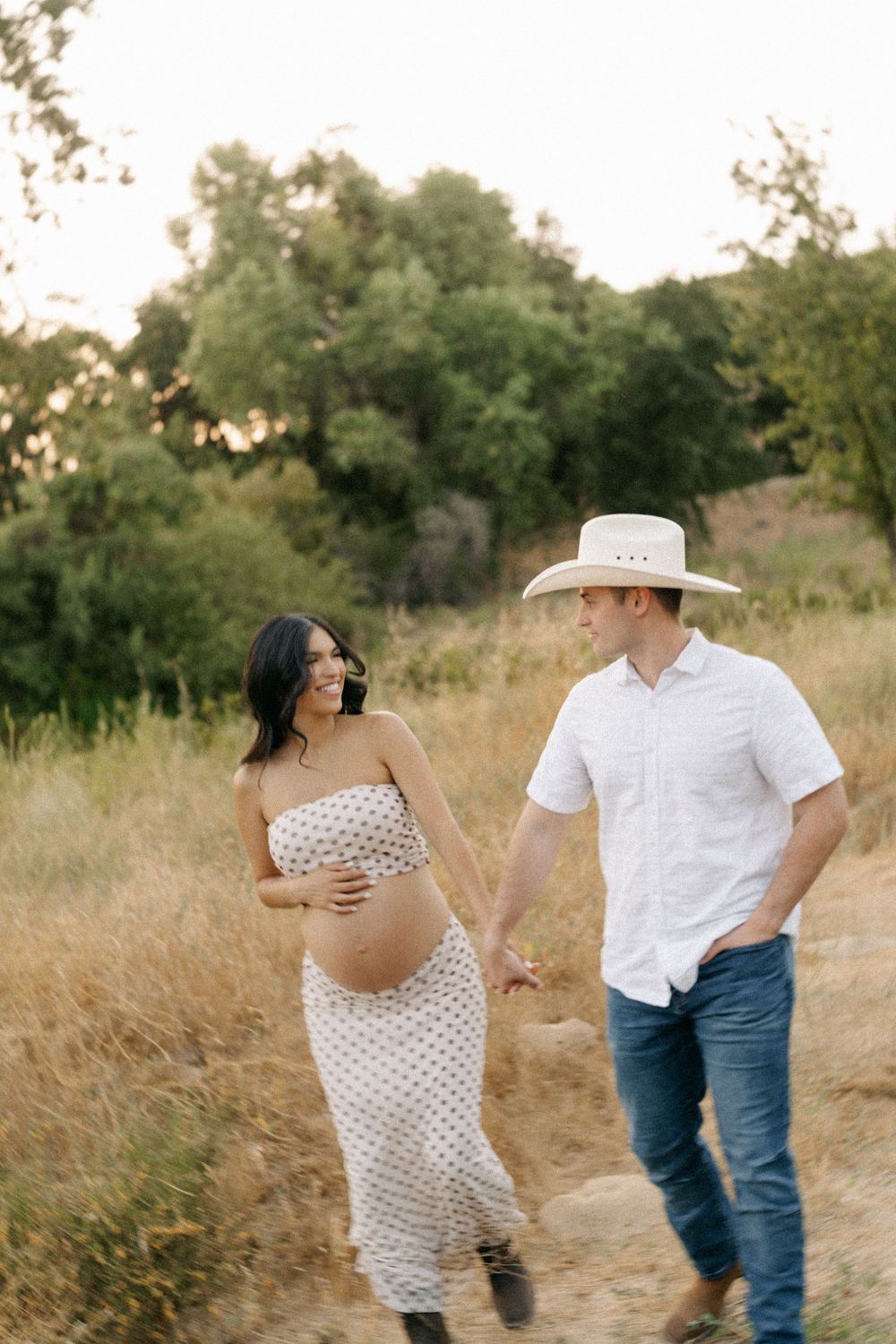 Pregnant couple walking hand in hand through golden field, woman in polka dot dress, man in white shirt and cowboy hat.