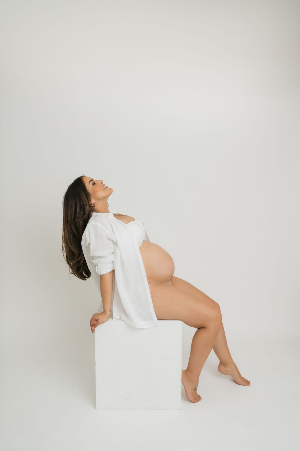 A minimalist maternity photoshoot featuring a person in a white button-down shirt against a light backdrop.